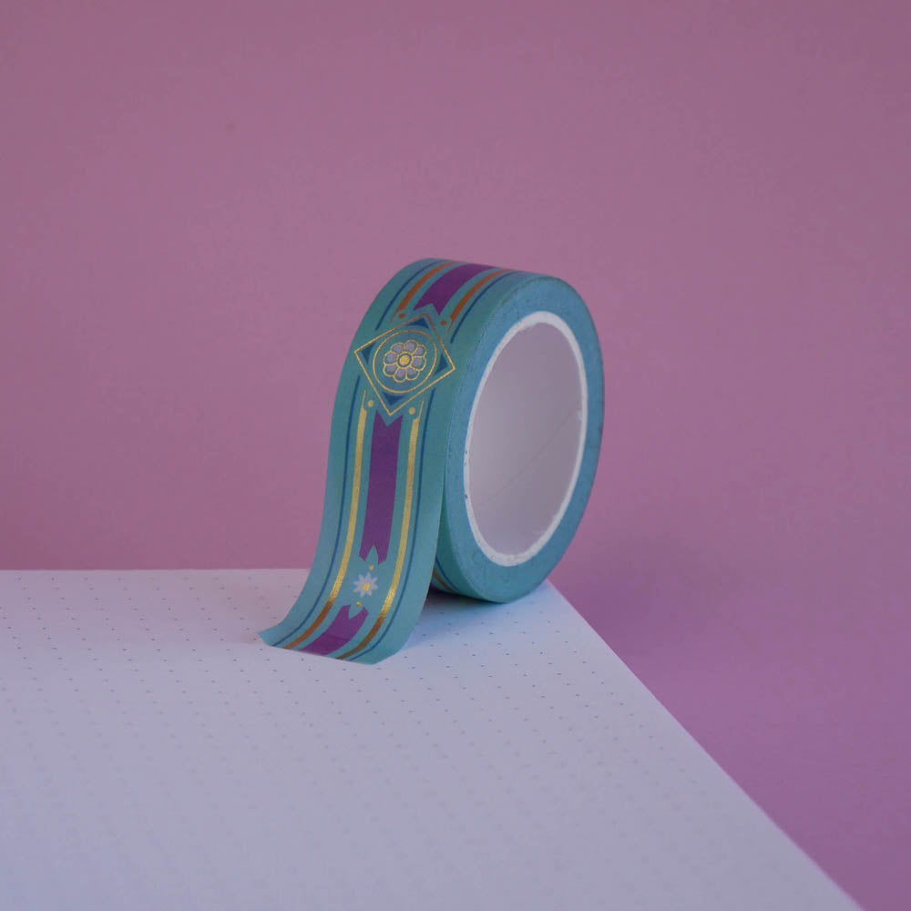 The Balteus design of French inspired washi tape as part of the Antoinette collection sits on a light pink background. The romantasy inspired washi tape features a classical book inspired motif in shades of blue, pink and foil gold.