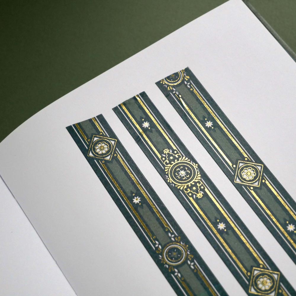 The Sage green variation of the Balteus Literati washi tape. The classic book inspired washi tape design is shown in three strips on the page of a journal sitting on a green background and features a classic victorian motif with a small flower in shades of dark green, sage green, white and foiled gold.