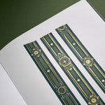 The Sage green variation of the Balteus Literati washi tape. The classic book inspired washi tape design is shown in three strips on the page of a journal sitting on a green background and features a classic victorian motif with a small flower in shades of dark green, sage green, white and foiled gold.