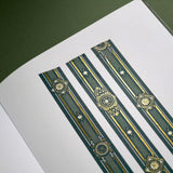 The Sage green variation of the Balteus Literati washi tape. The classic book inspired washi tape design is shown in three strips on the page of a journal sitting on a green background and features a classic victorian motif with a small flower in shades of dark green, sage green, white and foiled gold.