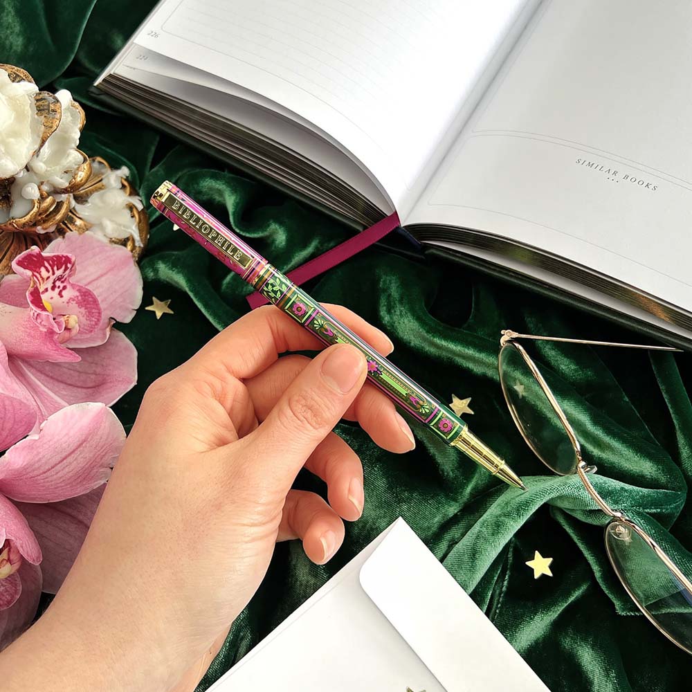 Hand holding a decorative pen with an open book, pink flowers, and glasses on a green surface.