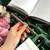 Hand holding a decorative pen with an open book, pink flowers, and glasses on a green surface.