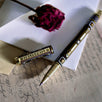 A hand holding the Black Bibliophile Pen by The Quirky Cup Collective, showcasing its intricate gold and white floral and geometric barrel design on a black background. The pen is positioned on top of a group of letters and envelopes with dried red roses creating a moody, vintage-inspired scene. 