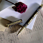 A hand holding the Black Bibliophile Pen by The Quirky Cup Collective, showcasing its intricate gold and white floral and geometric barrel design on a black background. The pen is positioned on top of a group of letters and envelopes with dried red roses creating a moody, vintage-inspired scene. 