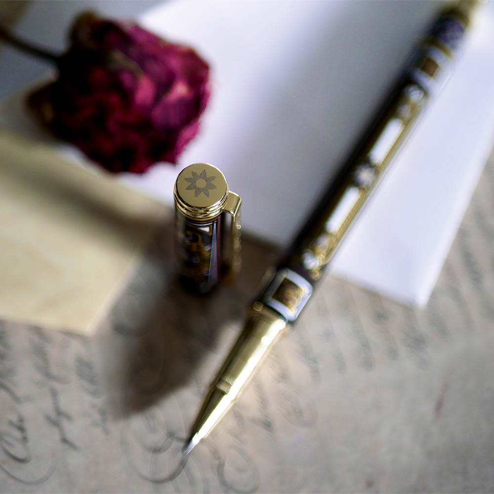 The Black Bibliophile Pen by The Quirky Cup Collective, showcasing its intricate gold and white floral and geometric barrel design on a black background. The pen is positioned on top of a group of letters and envelopes with dried red roses creating a moody, vintage-inspired scene. 