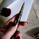 The Black Bibliophile Pen by The Quirky Cup Collective, showcasing its intricate gold and white floral and geometric barrel design on a black background. The pen is positioned on top of a group of letters and envelopes with dried red roses creating a moody, vintage-inspired scene. 