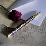 The Black Bibliophile Pen by The Quirky Cup Collective, showcasing its intricate gold and white floral and geometric barrel design on a black background. The pen is positioned on top of a group of letters and envelopes with dried red roses creating a moody, vintage-inspired scene. 