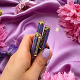A hand holds three decorative pens by The Quirky Cup Collective which feature an intricate victorian inspired design in shades of pastel pink and purple and the phrase 'Bibliophile' engraved on the clip. The pens are in front of a purple fabric background with flowers and small gold stars.
