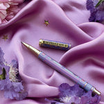 A decorative pen by The Quirky Cup Collective shows an intricate victorian inspired design in shades of pastel pink and purple and the phrase 'Bibliophile' engraved on the clip. The pen is on a purple fabric background with flowers and small gold stars.