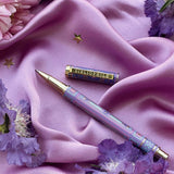 A decorative pen by The Quirky Cup Collective shows an intricate victorian inspired design in shades of pastel pink and purple and the phrase 'Bibliophile' engraved on the clip. The pen is on a purple fabric background with flowers and small gold stars.