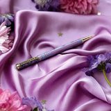 A decorative pen by The Quirky Cup Collective shows an intricate victorian inspired design in shades of pastel pink and purple and the phrase 'Bibliophile' engraved on the clip. The pen is on a purple fabric background with flowers and small gold stars.