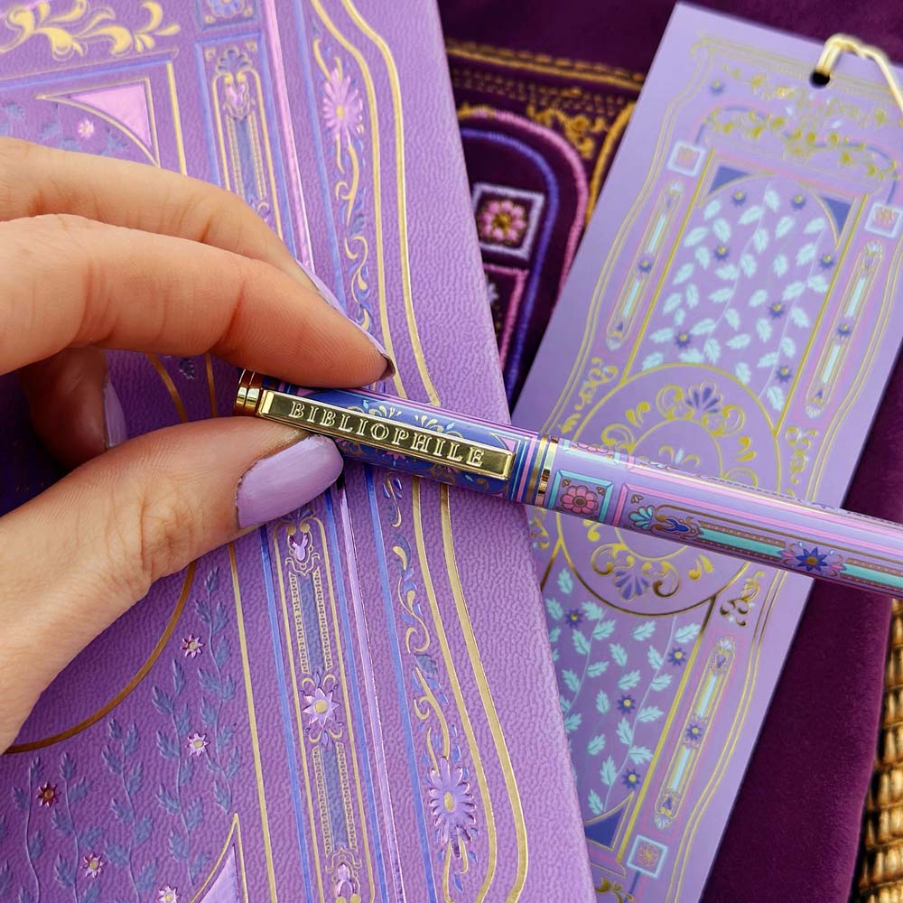 A decorative pen by The Quirky Cup Collective shows an intricate victorian inspired design in shades of pastel pink and purple and the phrase 'Bibliophile' engraved on the clip. The pen sits on a matching wisteria reading journal and bookmark.