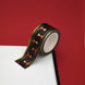 A close up of the Blood Spilled washi tape by The Quirky Cup Collective. The roll of fantasy inspired washi tape is on the corner of a white journal on a red background. The bookish washi tape design is red and black with gold detailing with a goblet motif.
