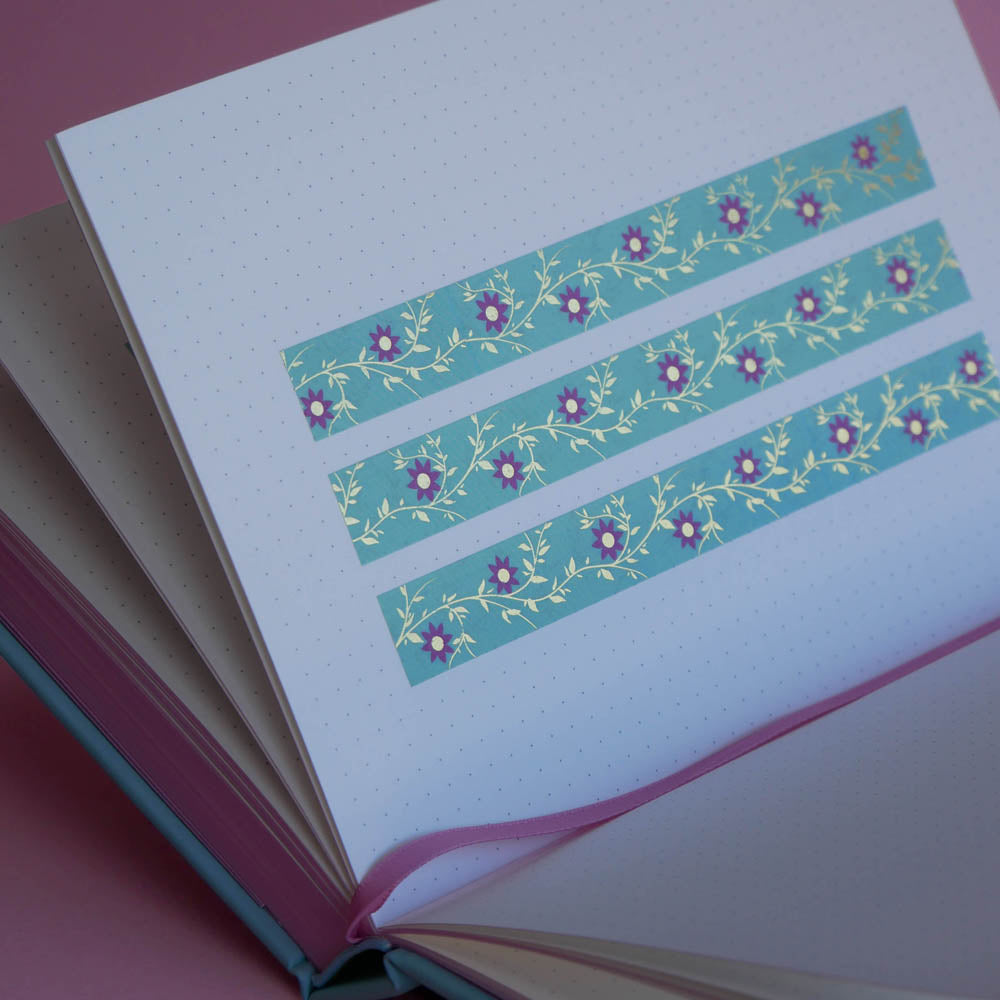 The Botanica washi tape in Antoinette features intricate flower and vine designs designs in shades of pink, gold and blue. Three stripes of the tape are in a page of the french inspired notebook against a pink background.