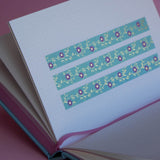 The Botanica washi tape in Antoinette features intricate flower and vine designs designs in shades of pink, gold and blue. Three stripes of the tape are in a page of the french inspired notebook against a pink background.