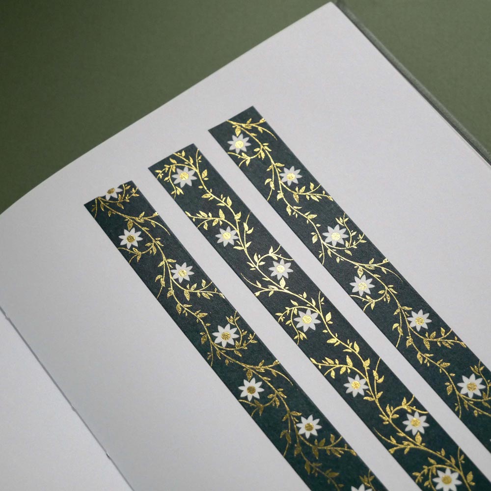 The Sage green variation of the Botanica Literati washi tape. The classic book inspired washi tape design is shown in three strips on a journal page on a green background and features a a vine covered in tiny flowers and leaves in shades of dark green, sage green, white and foiled gold.