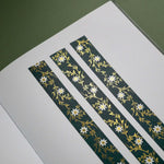 The Sage green variation of the Botanica Literati washi tape. The classic book inspired washi tape design is shown in three strips on a journal page on a green background and features a a vine covered in tiny flowers and leaves in shades of dark green, sage green, white and foiled gold.