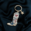 A denim coloured Cowboy boot keychain featured red and gold heart motifs and a small gold star sits on a deep green fabric background.