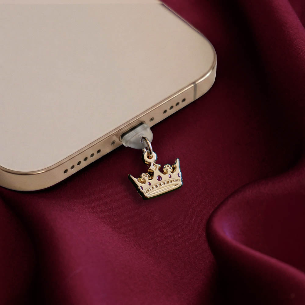 An detail shot of the gold Gilded Heir dust plug in a phone on a deep red fabric background. The fantasy aesthetic kindle decor sees the dust plug feature a small crown motif with red details.