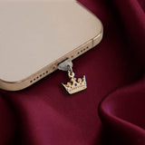 An detail shot of the gold Gilded Heir dust plug in a phone on a deep red fabric background. The fantasy aesthetic kindle decor sees the dust plug feature a small crown motif with red details.