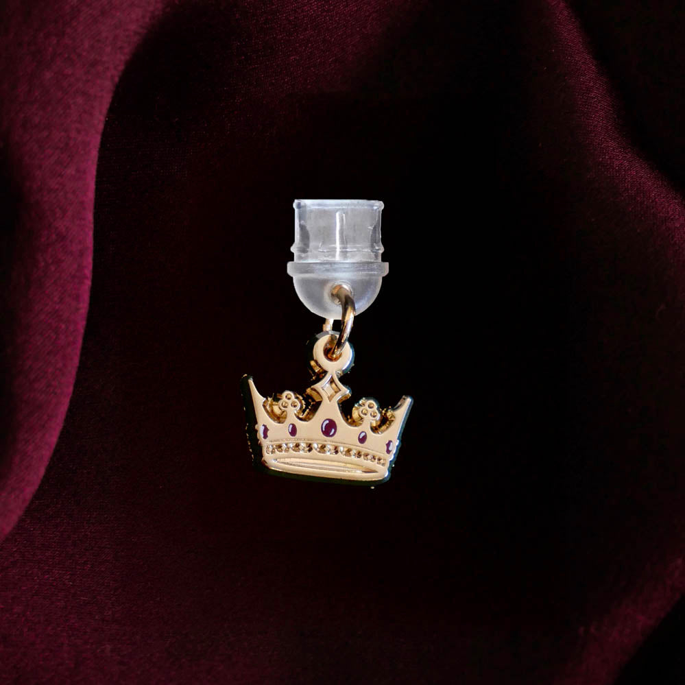 An detail shot of the gold Gilded Heir dust plug on a deep red fabric background. The fantasy aesthetic kindle decor sees the dust plug feature a small crown motif with red details.