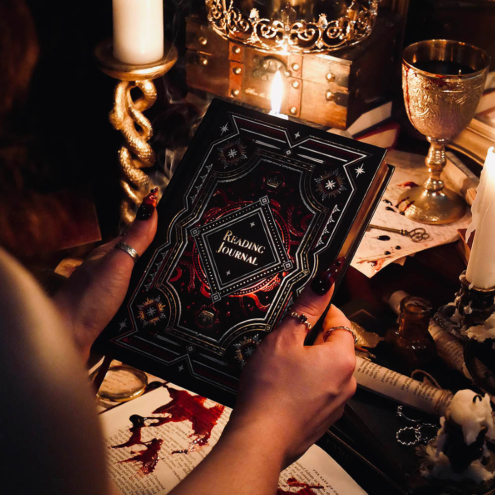 A promo image for the Serpents & Swords collection by The Quirky Cup Collective. A woman is holding a 'Reading Journal' with candles and a crown in the background. The journal is black with gold gilded pages and features red, gold and while foiling with snake and star motifs.