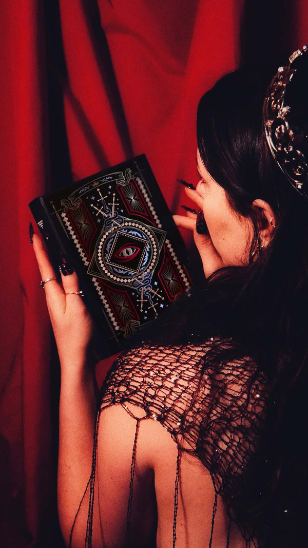 A woman wearing a crown holds an intricate journal by The Quirky Cup Collection against a red curtain backdrop. The foiled reading journal has a dragon eye motif alongside swords and ornate patterns.