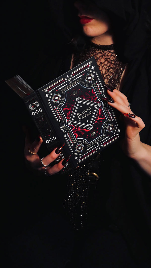 A woman wearing in a hood holds an intricate Reading Journal by The Quirky Cup Collection against a dark backdrop. The foiled reading journal has a crown motif alongside swords and snake patterns inline with the romantasy aesthetic.