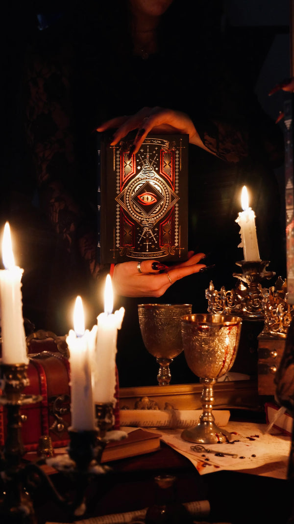 A promo image for the Serpents & Swords collection. Hands hold a lush reading companion in front of a mirror, candles and goblets surrounding the scene. The black journal features a dragon eye motif alongside swords and geometric patterns.