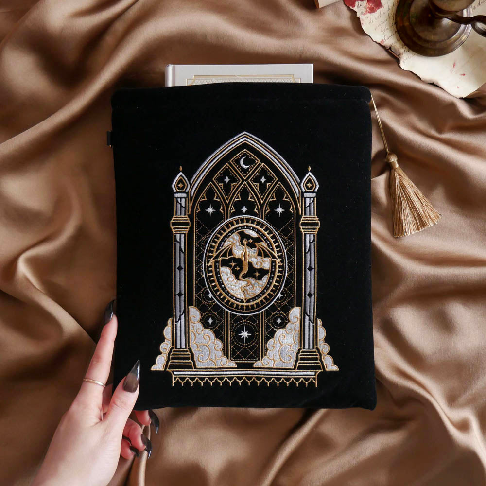The Dragon Lore embroidered book sleeve  sits on a warm gold fabric background. The black case features a gold tassel and a white, grey and gold romantasy inspired design of a dragon flying over a gothic stained glass window.