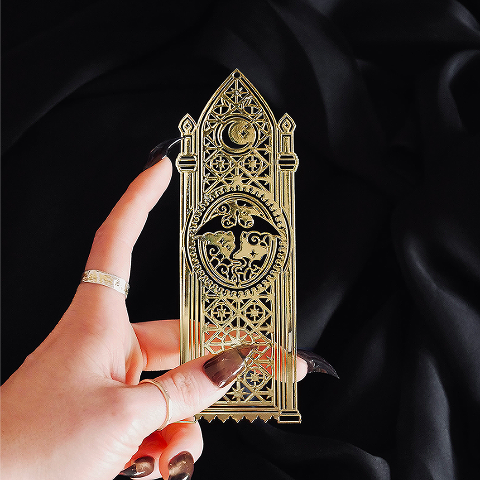 An intricate gold metal bookmark by The Quirky Cup Collective sits on a black background surrounded by candles and an open book. The bookmark features a stained glass inspired motif and has a flying dragon design at the center.
