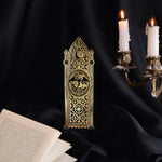 An intricate gold metal bookmark by The Quirky Cup Collective sits on a black background surrounded by candles and an open book. The bookmark features a stained glass inspired motif and has a flying dragon design at the center.