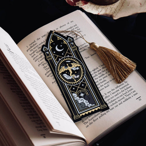 The Dragon Lore fantasy arch bookmark features gold and white foil detailing on a black background. The bookmark sits on an open classic book aesthetic and features a flying dragon motif with gothic archway design and a gold tassel.