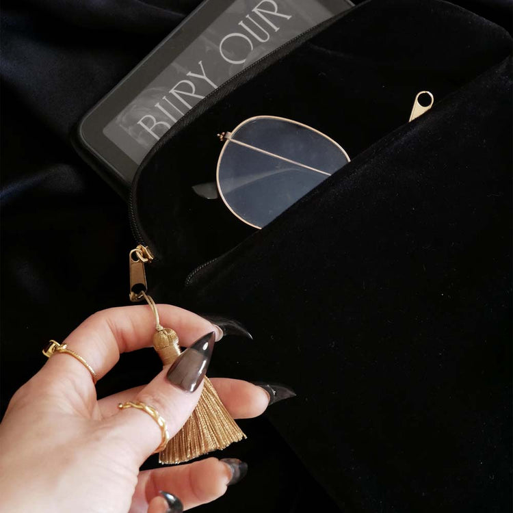 A detail of the back of Dragon Lore embroidered kindle case is held by a hand with long black nails on a black fabric background. The kindle case features a wrist strap and gold tassel. The black velvet kindle case features gold and cream embroidery of a dragon flying past an arched stained glass window through the clouds. 