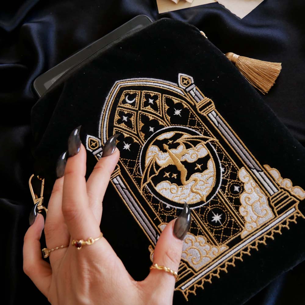 A detail of the back of Dragon Lore embroidered kindle case is held by a hand with long black nails on a black fabric background. The kindle case features a wrist strap and gold tassel. The black velvet kindle case features gold and cream embroidery of a dragon flying past an arched stained glass window through the clouds. 