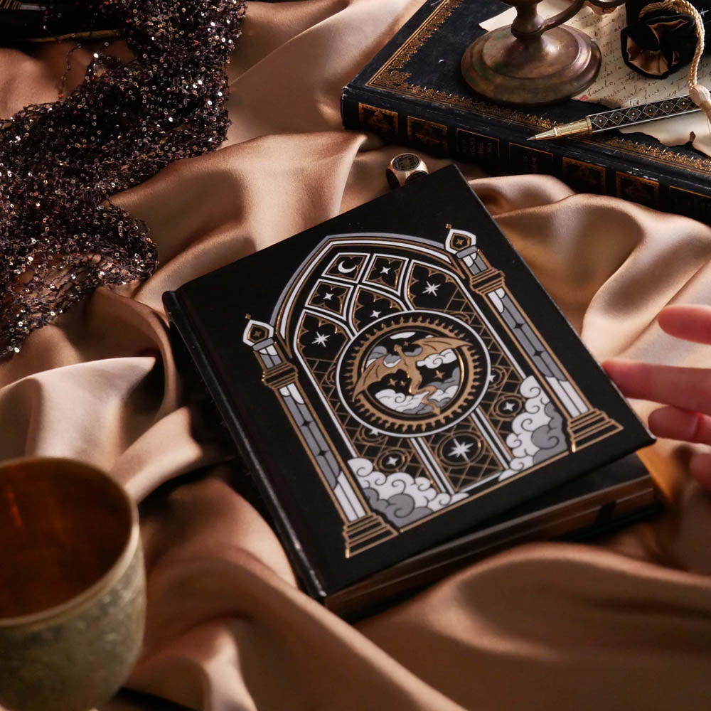The Dragon Lore journal sits on a beige fabric background surrounded by classic book aesthetic, candles and a matching romantasy pen. The journal features gold, silver and white foiling with a stained glass window design around a flying dragon.