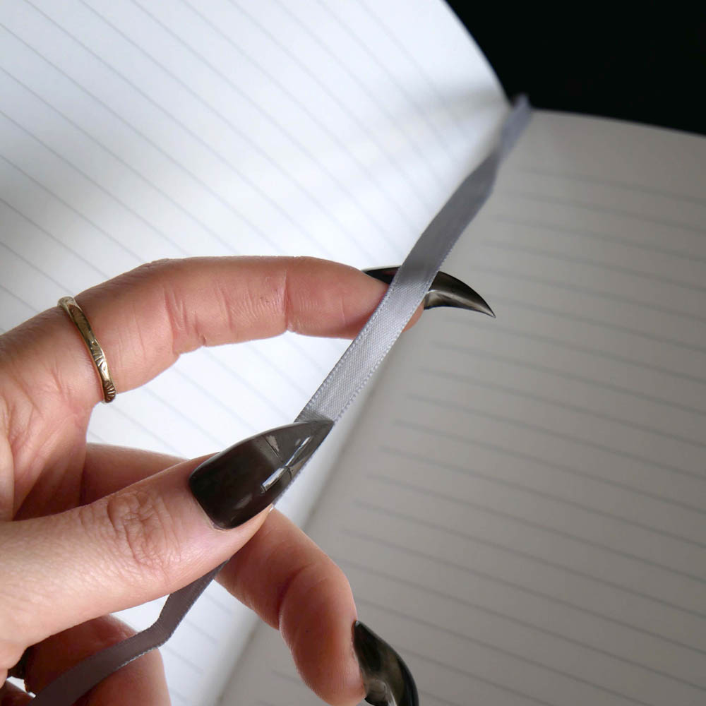 A close-up of a hand with dark brown nail polish and a gold ring holding a silver metal loop or clip against an open lined notebook.