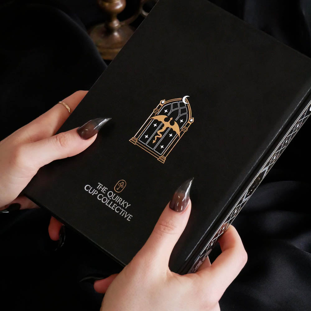 A person holds a black hardcover fantasy themed journal with a gold and black gothic emblem and the text 'THE QUIRKY CUP COLLECTIVE' on the cover. The person's hands with dark metallic nail polish are visible against a dark background.