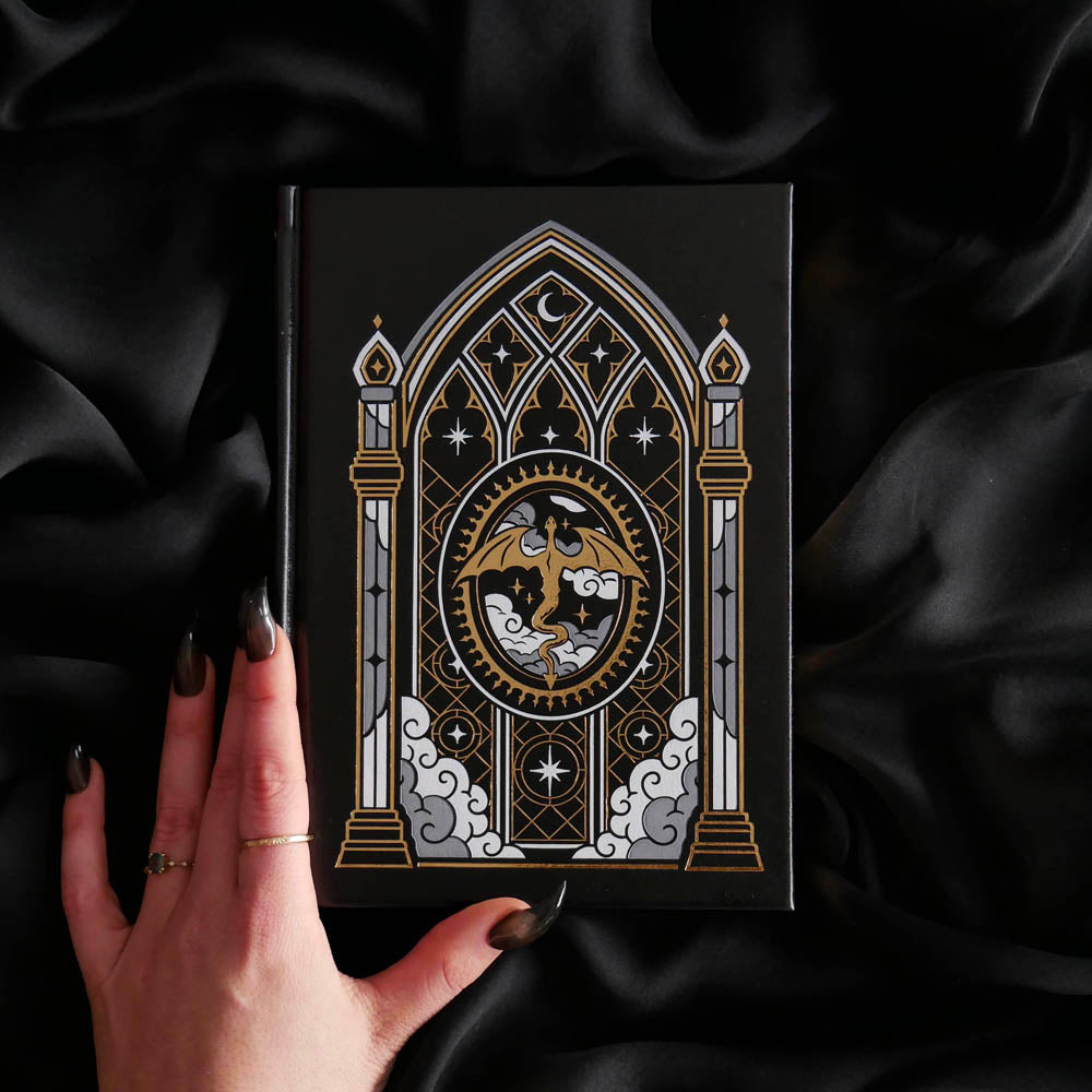 A black hardcover journal with an ornate gothic window illustration featuring a central dragon motif, held by a hand with dark manicured talons against a draped black satin background.