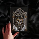 A black hardcover journal with an ornate gothic window illustration featuring a central dragon motif, held by a hand with dark manicured talons against a draped black satin background.