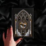 A black hardcover journal with an ornate gothic window illustration featuring a central dragon motif, held by a hand with dark manicured talons against a draped black satin background.