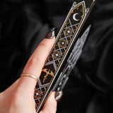 A close-up of a hand holding a black decorative fantasy dragon journal with gold and white cathedral window style symbols, including a crescent moon and stars, against a dark draped fabric background.
