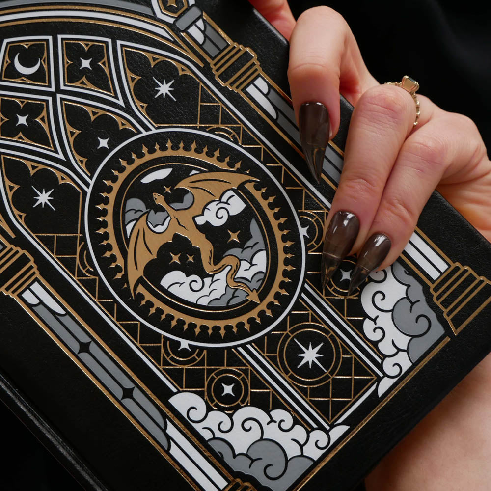 A close-up of a hand holding a black notebook or journal with an ornate Gothic-style dragon illustration in gold, gray, and white.
