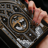 A close-up of a hand holding a black notebook or journal with an ornate Gothic-style dragon illustration in gold, gray, and white.
