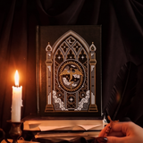 Fantasy book inspired A5 Faux leather black notebook with a celestial cathedral design. Centre of Journal features a dragon and cover has gold foil accents. Journal sits in an aesthetic setting that features a quill, dripping candle and messy bookstack. 