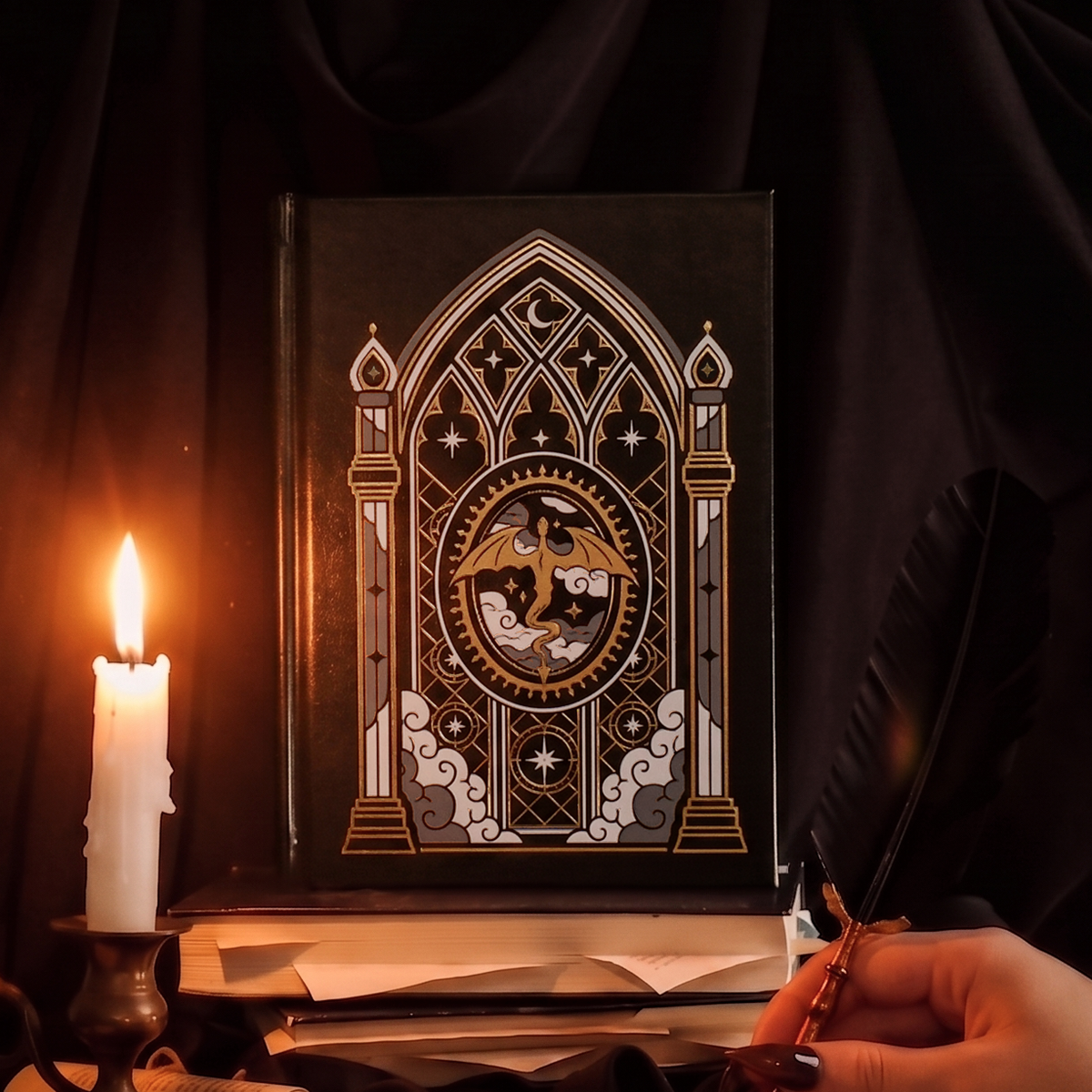 Fantasy book inspired A5 Faux leather black notebook with a celestial cathedral design. Centre of Journal features a dragon and cover has gold foil accents. Journal sits in an aesthetic setting that features a quill, dripping candle and messy bookstack. 