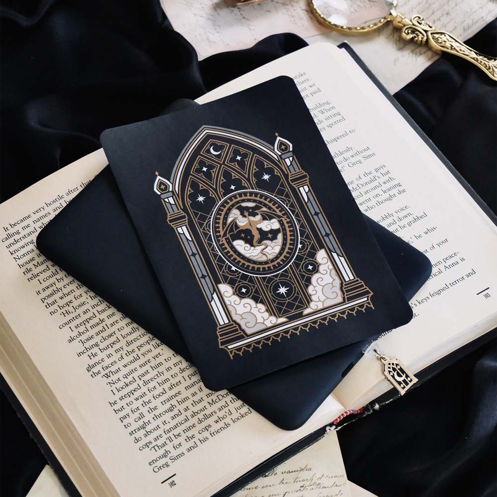 The Dragon Lore Kindle Sticker Insert by The Quirky Cup Collective is attached to a black kindle that sits on a classical book on a black fabric background. The black and gold sticker features a gold, cream and grey design of a dragon flying through clouds in front of a stained glass window. The kindle has a matching gold dust plug of the same arched window with moon and star motifs.
