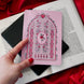 A hand holding a Kindle sticker insert over an e-reader on a background of a book and red fabric. The eReader sticker insert is light pink and features a stained glass window design with rose, plant and bird motifs in shades of red, pink and green.