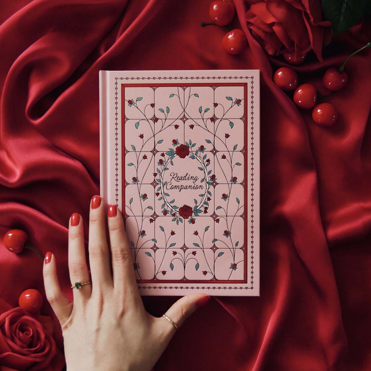 The Reading Companion - A pink journal laying across a draped red silk backdrop surrounded by roses and cherries. Pink Journal is inpspired by stained glass and includes roses and vines, centred with 'Reading Companion' text at the centre
