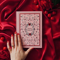 The Reading Companion - A pink journal laying across a draped red silk backdrop surrounded by roses and cherries. Pink Journal is inpspired by stained glass and includes roses and vines, centred with 'Reading Companion' text at the centre
